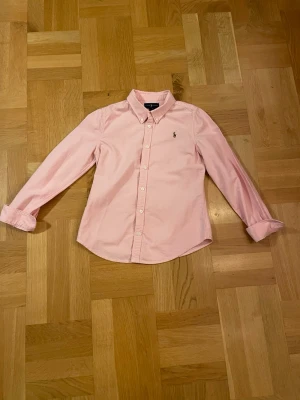 Rosa skjorta från Ralph Lauren - Snygg rosa skjorta från Ralph Lauren med klassisk krage och knappar framtill. Skjortan har lång ärm och ikonisk broderad logga på bröstet. Tillverkad i mjuk bomull som är skön mot huden. Perfekt för dig som gillar stilrena och tidlösa plagg.också väldigt bra om du vill preppa för sommarn.                                                                        det står storlek 12 och är lika med typ 146-152 tror jag efter som att jag är 158 och den börjar bli liten