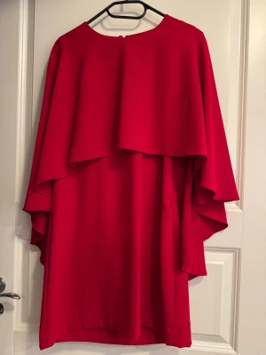 Röd omlottklänning från Zara - Säljer en röd omlottklänning från Zara med vid cape-liknande överdel och rund halsringning. Klänningen har en elegant och flytande siluett med kort längd och är tillverkad i ett mjukt syntetmaterial. Perfekt för dig som gillar statement-plagg.