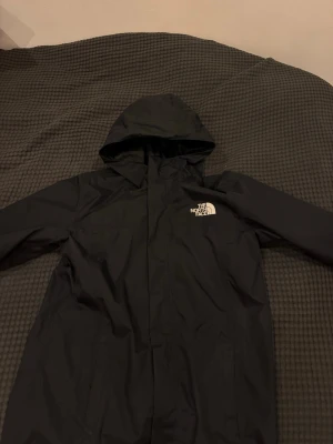 Svart vindjacka från The North Face - Säljer en svart vindjacka från The North Face med huva och meshfoder. Jackan har logga på bröstet och ärmen, samt elastiska muddar och dragkedja framtill. Perfekt för blåsiga dagar och enkel att packa ner i väskan. Det är storlek L/G alltså Large Grand i barn storlek 