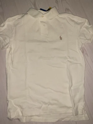 Vit pikétopp från Polo Ralph Lauren XS - Säljer en klassisk vit pikétopp från Polo Ralph Lauren i storlek XS. Modellen är Custom Slim Fit med korta ärmar, krage och knappar framtill. Toppens material är bomull och har den ikoniska broderade loggan på bröstet. Perfekt för en clean och stilren look. Använd 2 gånger 