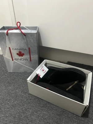 Svarta vinterskor/kängor från Canada Snow i svart - Säljer ett par svarta vinterskor/kängor från Canada Snow i svart med grov sula. Skorna har guldfärgade dragkedjor på sidan och en liten gulddetalj vid ankeln. Baktill finns en snygg logga på hällan. Perfekta för kalla dagar och riktigt stilrena. Undersidan är jätte snygg med röda detaljer och Canada loggan. Nypris 1800kr