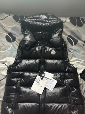 Svart dunväst från Moncler - Säljer en svart, glansig dunväst från Moncler med huva och två dragkedjeförsedda fickor. Västen har quiltad design och Moncler-logga på bröstet. Perfekt för kyliga dagar och har en modern, loose passform. Materialet är syntet och dun. Jag fick 2 på min födelsedag så jag valde och sälja en. (Kontakta vid minsta bekymmer)