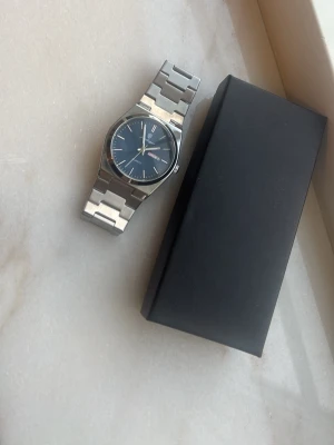 Klocka - Bilderna visar en klocka med metallarmband och svart urtavla, ingen justerings verktyg finns med.