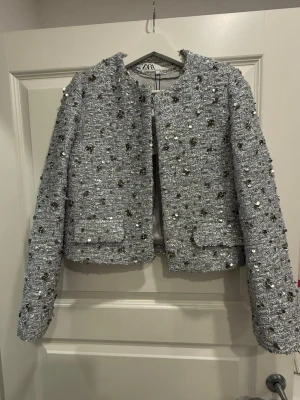 Glittrig silver kavaj från ZARA XS - Säljer en glittrig kavaj från ZARA i storlek XS. Den är silverfärgad med paljetter och blommiga detaljer. 