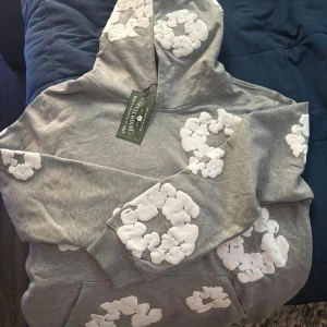 Grå hoodie med vita blommor från Denim Tears - Säljer en grå hoodie från Denim Tears med stora vita blommor tryckta över hela tröjan. Hoodien har huva, magficka och långa ärmar med ribbade muddar. Perfekt för dig som gillar streetwear och vill sticka ut med ett unikt mönster.