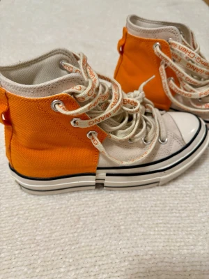 Converse x Feng Chen Wang orange sneakers - Unika höga sneakers från Converse i samarbete med Feng Chen Wang. Skorna har en cool split-sula, orange och beige canvas, rund tå och snörning med specialdesignade skosnören med logga. Perfekt för dig som vill sticka ut med din stil.