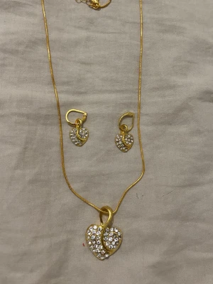 Fina halsband och örhängen  - Säljer ett smyckeset med halsband och örhängen i guldfärg. Både halsbandet och örhängena har hjärtformade hängen dekorerade med glittrande stenar. Kedjan är tunn och smidig, och hjärtana har en snygg böjd detalj i mitten. Perfekt för dig som gillar bling och hjärtan.