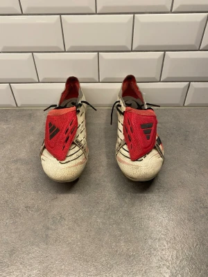 Adidas Predator fotbollsskor vit/röd - Säljer ett par Adidas Predator fotbollsskor i vitt med röda och svarta detaljer. Skorna har snörning, platt sula med dobbar och en röd tunga med Adidas-logga. Materialet är syntet och designen har svarta ränder på sidorna. Perfekt för dig som älskar fotboll och vill ha snygga skor på planen. Priset är inte hugget i sten