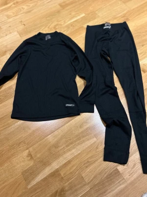 Svart underställ från Craft junior - Svart ribbat underställ från Craft i juniorstorlek 134/140. Setet består av långärmad tröja och byxor, tillverkade i mjuk polyester som andas. Perfekt för kalla dagar och träning utomhus. Diskret Craft-logga på både tröja och byxa.