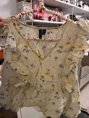 Blommig volangblus från vero moda - Supersöt blus med ljusgul bas och blommigt mönster i gult och lila. V-ringad framtill och volangdetaljer längs ärmar och nederkant. Perfekt för vår och sommar, luftig och lätt i bomull. Passar till jeans eller kjol för en fräsch look.