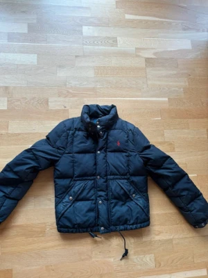 Svart pufferjacka från Polo Ralph Lauren - Svart pufferjacka från Polo Ralph Lauren med röd logga på bröstet. Jackan har hög krage, dragkedja och knappar framtill, samt två stora fickor med knappstängning. Justerbar snörning nedtill och quiltad design för extra värme.