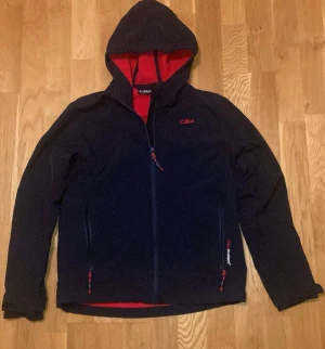 Svart softshell jacka med röd insida från CMP - Svart softshell från CMP med röd insida och huva. Jackan har dragkedja framtill, två sidofickor med röda detaljer och reflextryck på ryggen. Perfekt för blåsiga och regniga dagar och har en sportig look som funkar till många outfits.