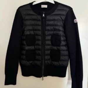 Moncler cardigan  - Svart pufferjacka från Moncler med stickade detaljer på framsidan och ärmarna. Jackan har en rund hals, dragkedja framtill och Moncler-logga på ena ärmen. Kombinerar quiltad pufferstil med stickade partier för en cool look. Taggar finns kvar! 