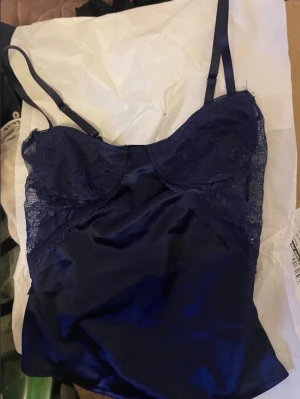 Mörkblå satin korsett med spets - Säljer en mörkblå korsett-topp i glansig satin med vackra spetsdetaljer över bysten och sidorna. Toppens axelband är smala och justerbara för perfekt passform. Perfekt för dig som vill ha en elegant och stilren look.