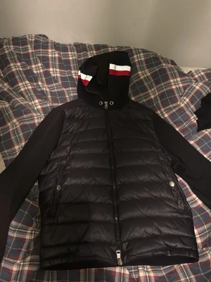 Svart Moncler cardigan med reflex luva - Svart cardigan från Moncler med quiltad front och släta ärmar. Jackan har en huva med vita, röda ränder och även reflex i mitten, dragkedja framtill och två sidofickor med metallknappar. Moncler-logga på ärmen ger extra premiumkänsla. Perfekt för dig som gillar stil och komfort.