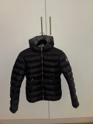 Svart dunjacka från Colmar med huva - Säljer en svart dunjacka från Colmar med huva och två fickor med dragkedja. Jackan har en quiltad design och är tillverkad i polyester. Perfekt för kalla dagar och har en snygg, sportig look. Passar dig som gillar stilrena och funktionella jackor.