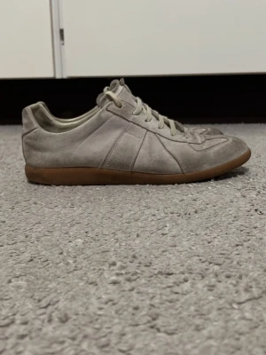 Margiela gats beige  - Säljer ett par beige sneakers från Maison Margiela i mjuk mocka med klassisk gummisula. Skorna har snörning och en stilren, retroinspirerad design med rund tå. Perfekta för dig som gillar minimalistisk stil och neutrala färger.