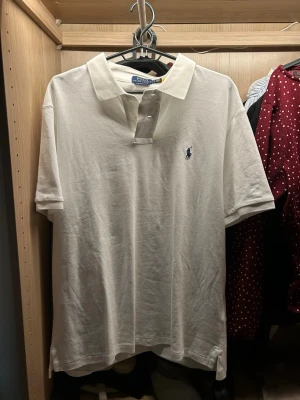 Vit polotopp från Polo Ralph Lauren - Säljer en klassisk vit polotopp från Polo Ralph Lauren med korta ärmar och krage. Toppen har knappar vid halsen och den ikoniska broderade loggan i marinblått på bröstet. Perfekt för en clean och stilren look.