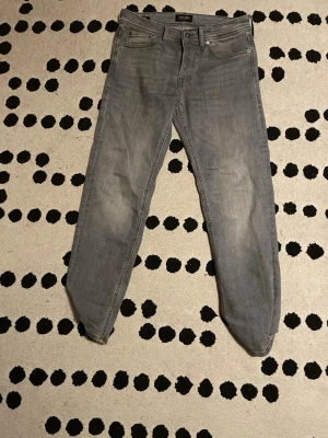 Grå tapered jeans från Jack & Jones - Snygga grå jeans från Jack & Jones, modell Tapered Mike. Jeansen har klassisk femficksdesign, smalare ben och normal midja. Materialet är mjukt denim i bomull med en lätt tvättad look. Perfekt för dig som gillar stilrena och bekväma jeans.