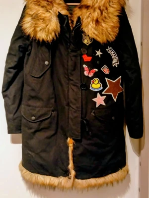 Svart parkajacka med päls från Kengstar - Parkajacka i svart med stora bruna fuskpälsdetaljer vid krage och nederkant. Jackan har coola broderade patches med stjärnor, fjäril, giraff och andra motiv både fram och bak. Flera fickor med knappar och dragkedja framtill. Perfekt för dig som gillar unika detaljer.