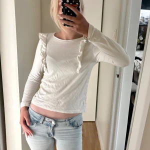 Vit långärmad topp med volang Hampton Republic - Säljer en vit långärmad topp från Hampton Republic i mjuk bomull. Toppen har volangdetaljer längs axlarna och en rund halsringning. Passar dig som gillar stilrena plagg med en liten twist. Perfekt att matcha med jeans eller kjol. Passar xs