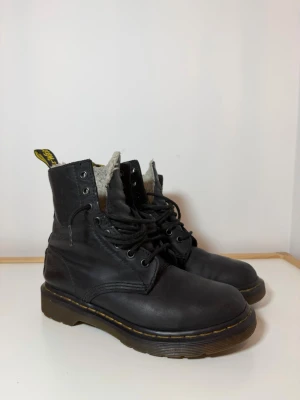 Svarta Dr. Martens boots med foder - Säljer ett par svarta Dr. Martens boots med rund tå och snörning. Skorna har en klassisk gul söm runt sulan och är fodrade med mjukt material för extra värme. Tillverkade i skinn med robust platt sula och tydliga dragflikar med gul text baktill.
