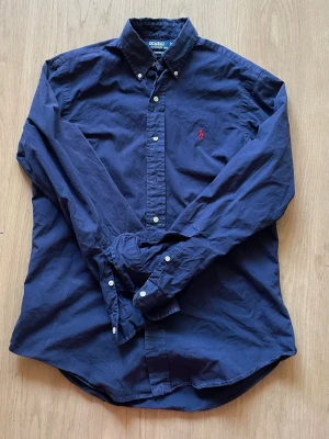 Marinblå skjorta från Ralph Lauren - Snygg marinblå skjorta från Ralph Lauren med klassisk röd broderad logga på bröstet. Skjortan är i storlek M och är i mycket gott skick dock saknas en knapp.