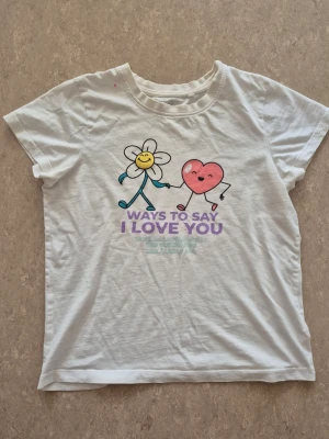 T-shirt - Vit t-shirt från Wow i mjuk bomull med korta ärmar och rund hals. Framsidan har ett färgglatt tryck med en glad blomma och ett hjärta som håller hand, samt texten 'Ways to say I love you' i lila. Perfekt för dig som gillar gulliga motiv och sköna basplagg. Finns en rosa fläck.
