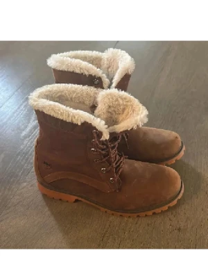 Bruna Helly Hansen boots med foder - Säljer ett par bruna boots från Helly Hansen med mjukt, ljust foder och grov sula. Skorna har snörning och är tillverkade i mocka med slitstark gummisula. Perfekta för kalla dagar och har en klassisk outdoor-look.