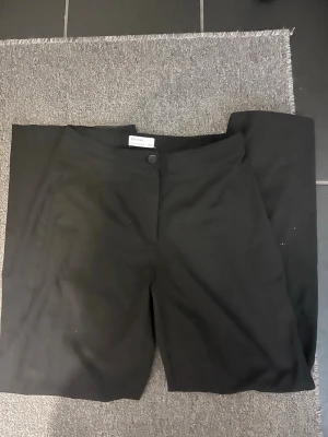 Svarta kostymbyxor från Bershka - Snygga svarta kostymbyxor från Bershka i klassisk rak modell. Byxorna har knapp och dragkedja framtill samt diskreta sidfickor. Perfekta för en clean och stilren look. Materialet är mjukt och har en lätt stretch för extra komfort.