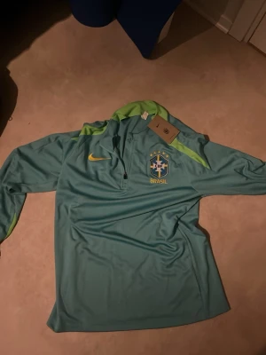 Nike Brasil träningströja blågrön - Nike Brasil träningströja i blågrön färg med limegröna detaljer och halv dragkedja. Broderat Brasilien-emblem med fem stjärnor på bröstet och gul Nike-logga. Tillverkad i polyester, perfekt för fotbollsträning.