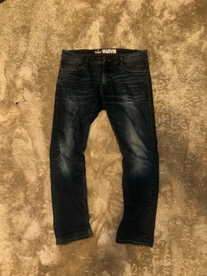 Mörkblå Marvin jeans straight fit - Snygga mörkblå jeans från Marvin med straight fit och klassisk femficksdesign. Jeansen har diskreta slitningar och kontrastsömmar, samt detaljerade bakfickor med broderade mönster. Perfekta för dig som gillar en enkel och stilren look.