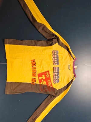 Gul långärmad tröja från D&G - Cool långärmad tröja från D&G i gul och brun med lila krage. Tryck på framsidan med texten 'GOOD IDEAS', 'FOLLOW ME' och 'MY FAV T-SHIRT'. Materialet är bomull och polyester, vilket gör den skön och lätt att bära. Perfekt för dig som gillar färgstarka och unika plagg. Storleken motsvarar Medium.