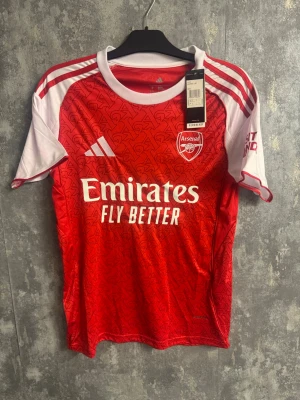 Arsenal matchtröja - Ny storlek S - Säljer en officiell Arsenal matchtröja från Adidas i rött och vitt med snyggt mönster och klubbmärke på bröstet. Tröjan har vita axelpartier med röda ränder och korta ärmar. Materialet är lätt polyester och perfekt för fotboll eller träning.