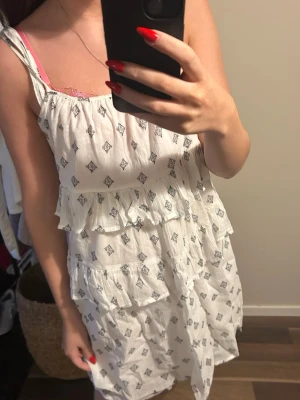 Vit kortklänning med volanger - Säljer en söt vit kortklänning med svarta små mönster och volangdetaljer. Klänningen har ärmlös design med smala axelband och ett luftigt, flowy tyg som känns perfekt för varma dagar. Passformen är loose och materialet känns som bomull.