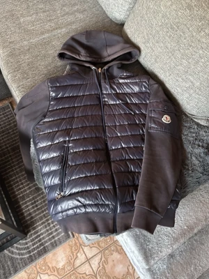 Moncler mörkblå pufferjacka med huva - Mörkblå cardigan från Moncler med huva och ribbade muddar. Jackan har quiltad framsida i glansigt material, släta ärmar och en ficka med Moncler-logga på armen. Dragkedja framtill och två sidofickor med zip. Perfekt för dig som vill ha en snygg och sportig look. Ena dragtjedjan e sönder men går att fixa med lite tid. Lite solblekt ochså men inget man tänker på.