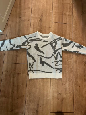 Vit och grå stickad tröja med mönster - Säljer en stickad tröja i vit bas med grått abstrakt mönster. Tröjan har rund hals och långa ärmar, samt ribbade muddar vid hals, ärmslut och nederkant. Perfekt för dig som gillar unika detaljer och sköna material. Helt ny, oanvänd