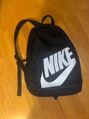 Svart ryggsäck från Nike - Säljer en svart ryggsäck från Nike med stort vitt logotryck på framsidan. Väskan har justerbara axelremmar, ett huvudfack med dragkedja och en mindre ficka framtill. Perfekt för skola eller träning, stilren och enkel att matcha.