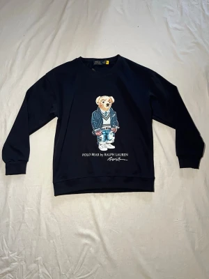 Mörkblå Tröja Polo Bear Ralph Lauren - Svart tröja från Polo Ralph Lauren med ikoniska Polo Bear-trycket framtill. Tröjan har rund hals, långa ärmar och ribbade muddar. Tillverkad i mjuk bomull som är skön att bära. Perfekt för dig som vill ha en klassisk men lekfull look. Storlek M men passar bättre för S. Kontakta oss vid minsta fråga⭐️✅