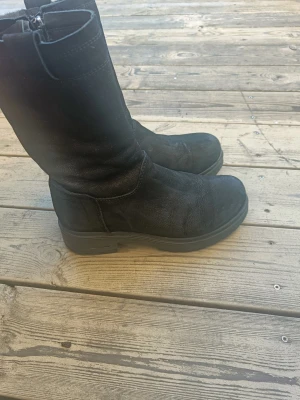 Svarta boots i skinn med dragkedja - Säljer ett par svarta boots i skinn med grov platt sula och rund tå. De har dragkedja på sidan för enkel på- och avtagning. Perfekta för dig som gillar en stilren och robust look. Passar till både jeans och kjol.