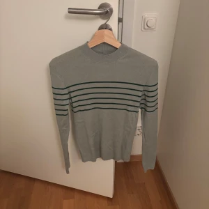 Grå randig tröja Massimo Dutti M - Snygg grå långärmad tröja från Massimo Dutti i storlek M. Tröjan har mörkgröna horisontella ränder över bröstet och armarna, ribbad struktur och rund hals. Perfekt för dig som gillar stilrena och klassiska plagg med en twist.