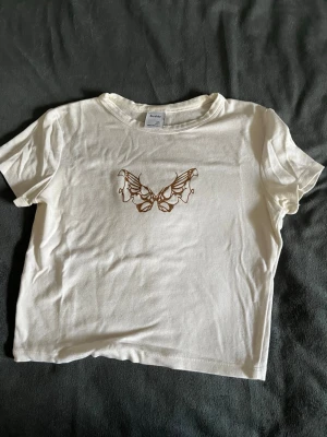 Vit t-shirt med fjäril från Bershka - Säljer en vit t-shirt från Bershka i storlek S. T-shirten har ett snyggt tryck med en brun fjäril på bröstet och klassisk rund hals. Modellen är kortärmad och gjord i mjuk bomull.