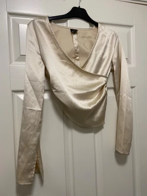 Champagnefärgad omlottopp Gina Tricot - Säljer en champagnefärgad omlottopp från Gina Tricot i storlek XS. Toppen har lång ärm, v-ringad hals och är tillverkad i glansigt satinmaterial. Snygg drapering och rynkade detaljer på sidan ger en cool look. Perfekt att styla med jeans eller kjol. Endast använd en gång, så väldigt fint skick.