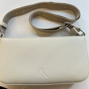 Vit/beige axelväska från Calvin Klein - Snygg vit/beige axelväska från Calvin Klein med präglad CK-logga framtill och bred axelrem med broderad logotyp. Väskan är tillverkad i skinnimitation med struktur och har både innerfack med dragkedja och ett praktiskt ytterfack. Perfekt storlek för det viktigaste. Den är inte längre i originalförpackning men har aldrig använts 
