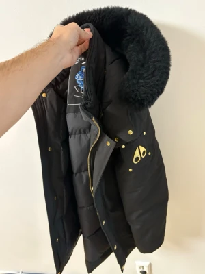 Svart pufferjacka Moose Knuckles - Säljer en svart pufferjacka från Moose Knuckles med fluffig päls runt kragen och guldfärgade detaljer. Jackan har broderad logga på insidan och metallmärke på ärmen. Fodret är quiltat och jackan stängs med dragkedja och knappar.