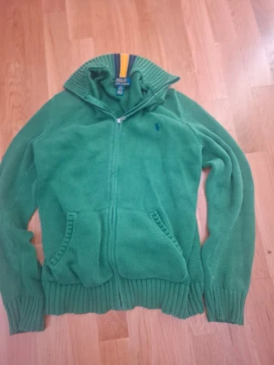Grön stickad hoodie från Polo Ralph Lauren - Säljer en grön stickad hoodie från Polo Ralph Lauren, äkt från Kids brand store med dragkedja framtill och två fickor. Tröjan har ribbade muddar och krage med snygga ränder i blått och gult. Perfekt för dig som gillar klassisk streetstil och vill ha något bekvämt och snyggt.