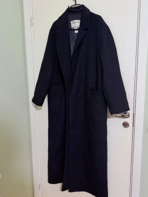 Svart lång kappa från Brixtol Textiles - Säljer en svart, stilren och lång kappa från Brixtol Textiles i storlek L. Kappan har klassisk krage, dubbla knappar framtill och är tillverkad av högkvalitativ ull med återvunna fibrer. Perfekt för dig som gillar minimalistisk och tidlös design.