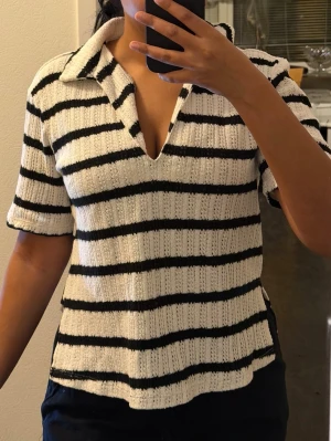 Randig stickad polotopp med krage - Säljer en vit och svart randig stickad polotopp med korta ärmar och djup v-ringning. Toppen har en bred krage och ribbad struktur som ger en snygg och avslappnad look. Perfekt att styla med jeans eller kjol.