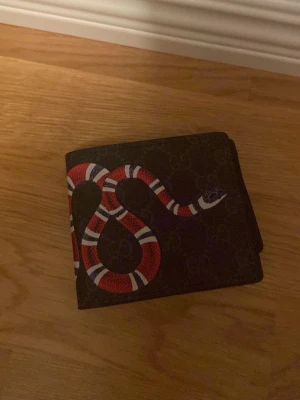 Svart Gucci plånbok med orm - Säljer en svart plånbok från Gucci med ett coolt ormmotiv i rött, vitt och blått på framsidan. Plånboken är tillverkad i skinn och har flera fack för kort. Perfekt för dig som vill ha en unik accessoar med lyxig känsla.