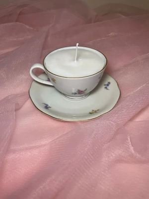 Doftljus i tekopp i en vit färg med flerfärgat blommigt mönster  - Det här ljuset är gjort av återanvändbara material. Tekoppen är köpt secondhand och stearinet är från kyrkor som tänkte slänga det. Det här ljuset har en svag doft utav vanilj, så känsliga även kan använda det.